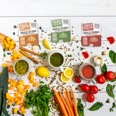 Vibrant vegetables scattered across a white wood plank background, with three mugs containing soup. Packets of Wild Zora New Vegan Soups are placed above the veggies and mugs of soup. 